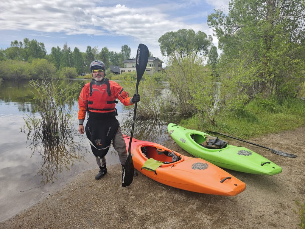 Tom holding a paddle, standing by two kayaks, at the edge of the water.
